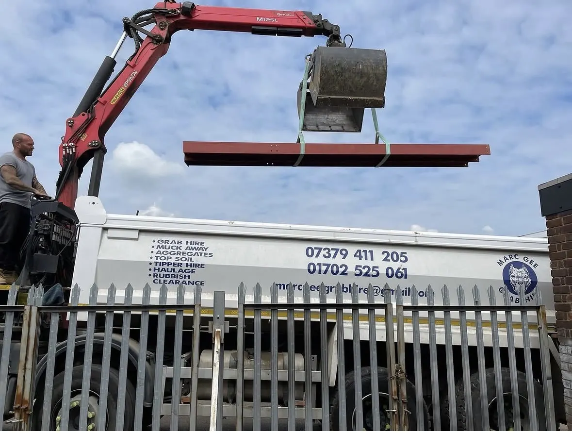 Grab crane lifting red steel beams in a yard, operator at the controls