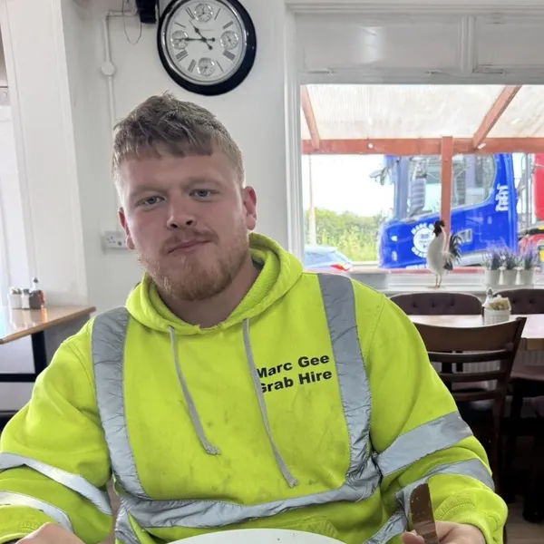 Logan in a Marc Gee Grab Hire hi-vis hoodie with a fry-up, a Marc Gee lorry parked outside the window
