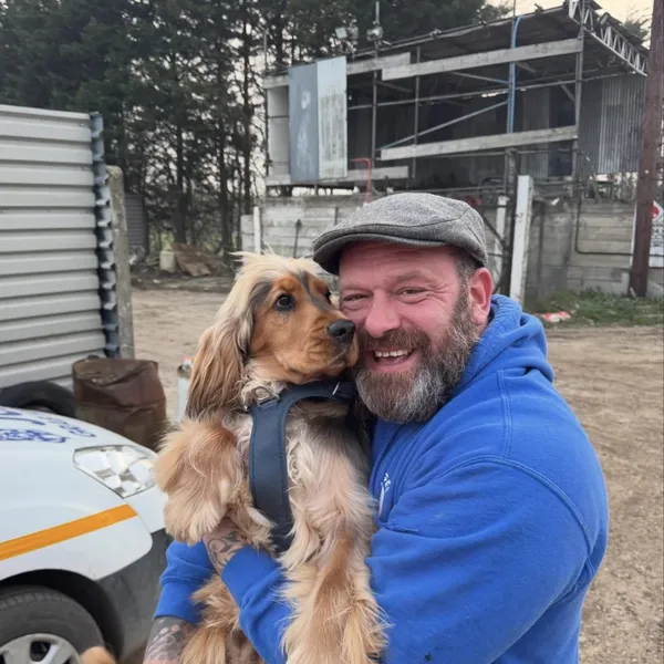 Marc Gee in a flat cap and blue hoodie, cuddling a spaniel in the yard